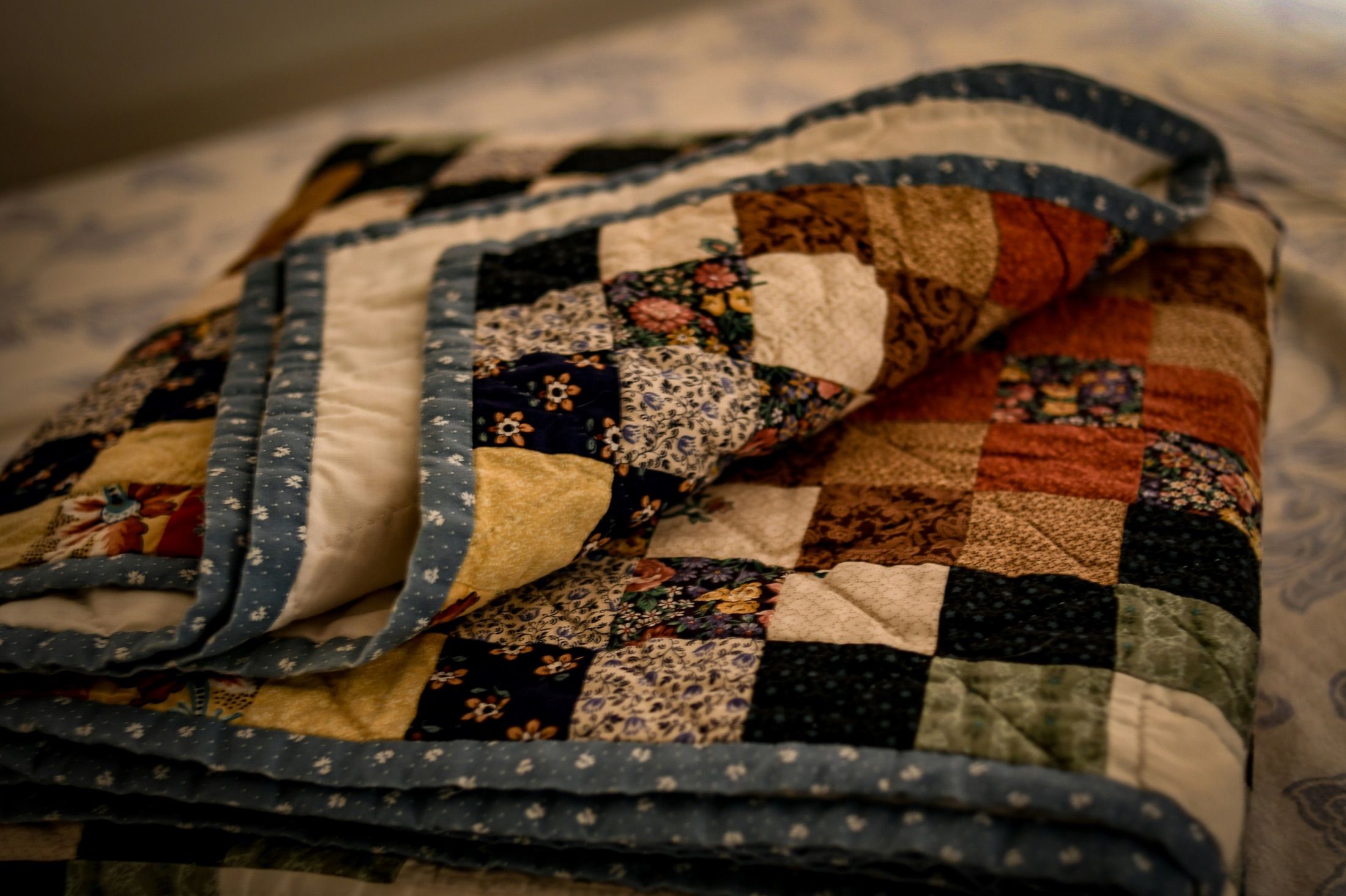 A folded patchwork quilt with a blue patterned binding resting on a bed